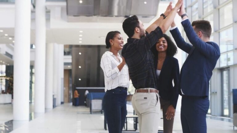 Grupa zaposlenih u poslovnom prostoru slavi uspeh i daje “high five”, što simbolizuje timski duh i zajedništvo. Fotografija prikazuje pozitivnu atmosferu na tim building događaju ili konferenciji.